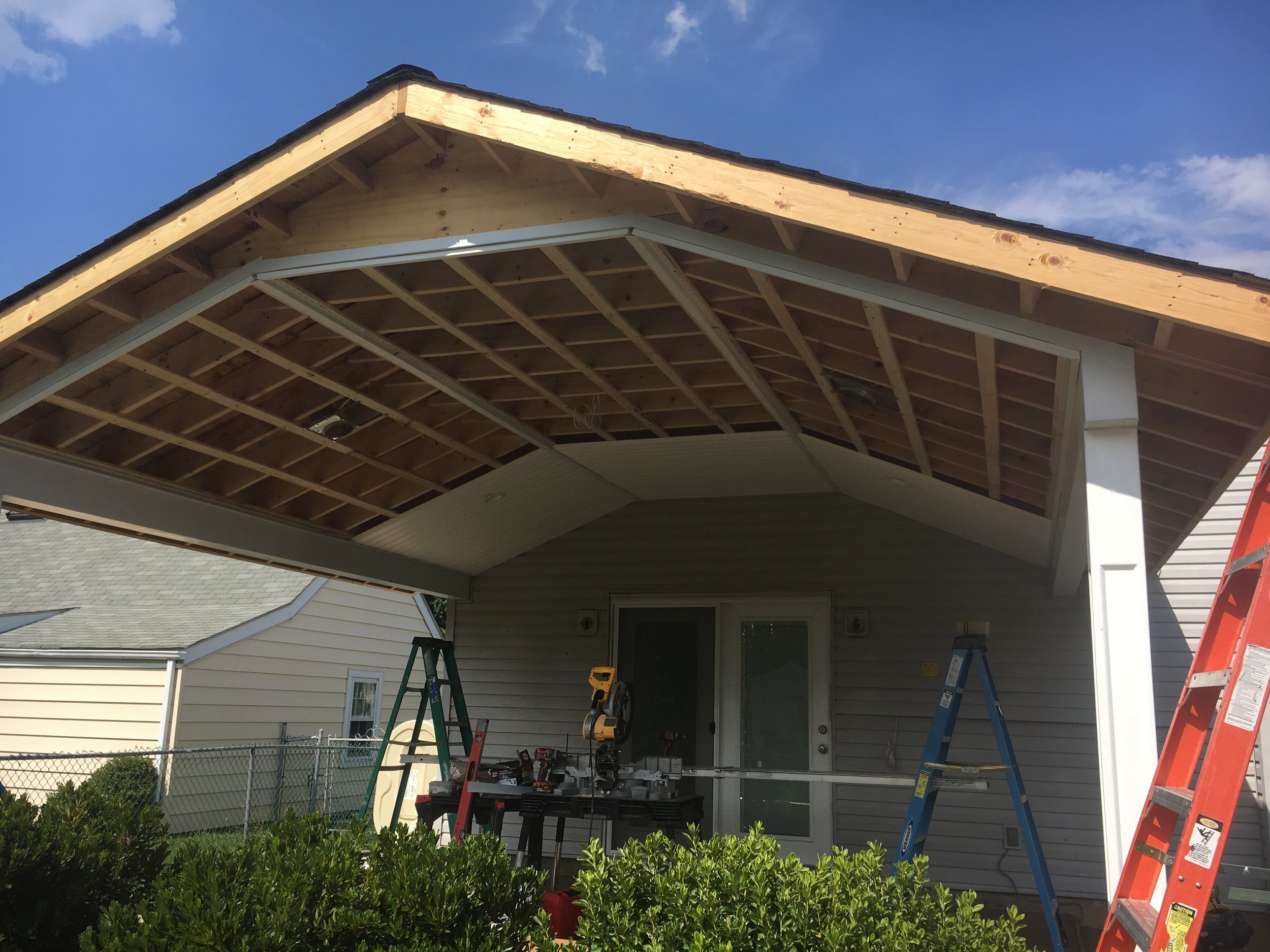 Under construction porch or patio cover with wooden framing and tools, ladders, and bushes in front.