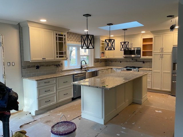 Kitchen with white cabinets, granite countertops, island, stainless steel appliances, pendant lights, skylight, and backsplash tiles, under renovation.