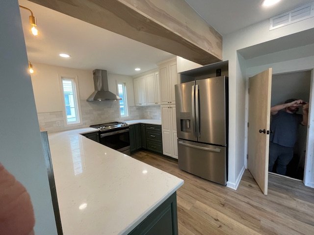 Modern kitchen with white upper cabinets, dark green lower cabinets, stainless steel refrigerator, and a stainless steel range with hood. Light wood flooring and a small doorway leading to a room with a person taking a photo.