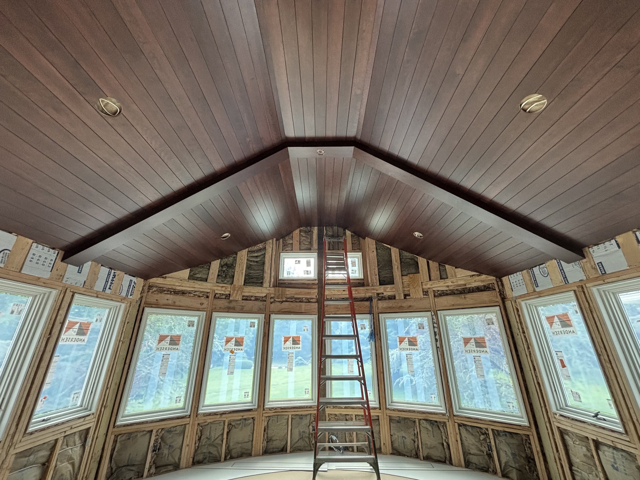 Interior of a house under construction with a partially finished wooden ceiling, multiple windows, a ladder in the center, and insulation visible around the walls.