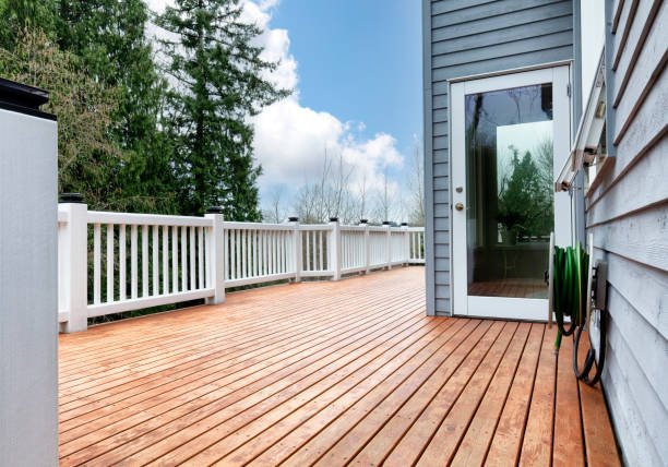 Empty outdoor wooden deck with white railing, adjacent to a blue house with a glass door, under a partly cloudy sky with trees in the background.