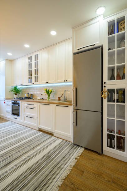 Modern kitchen with white cabinets, stainless steel refrigerator, built-in oven, and glass-fronted wine storage cabinets, with a striped rug on wooden floor.