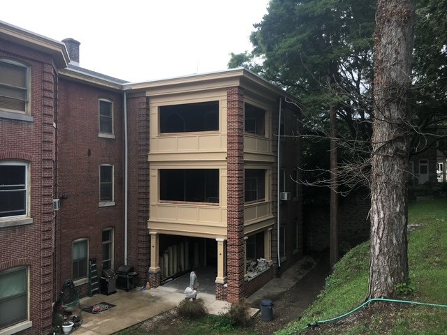 Three-story residential building with beige balconies and red brick exterior, surrounded by trees and a small yard with various items and black trash bins.