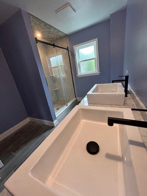 Modern bathroom with a walk-in shower with glass door, double sinks with black faucets, and a window on a blue wall.