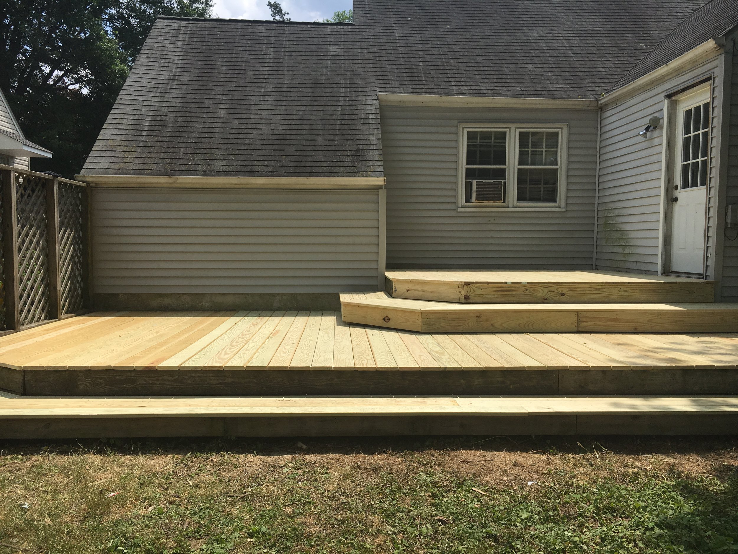 Newly built wooden deck with stairs in backyard, attached to house with siding exterior, double-hung windows, and a white door, with a grassy yard in the foreground.
