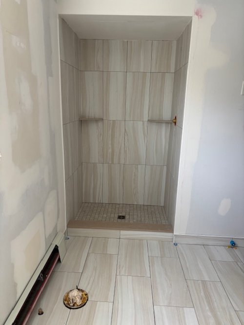 Empty tiled shower stall with two small built-in shelves. The shower is unfinished, with visible drywall and a floor drain. There is a small round object with a pattern on the floor in front of the shower.