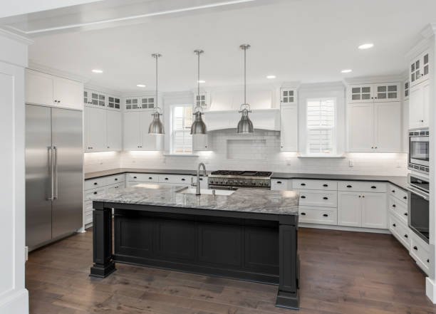 Modern kitchen with white cabinets, stainless steel refrigerator, built-in oven, gray kitchen island with marble countertop, pendant lighting, and hardwood floors.