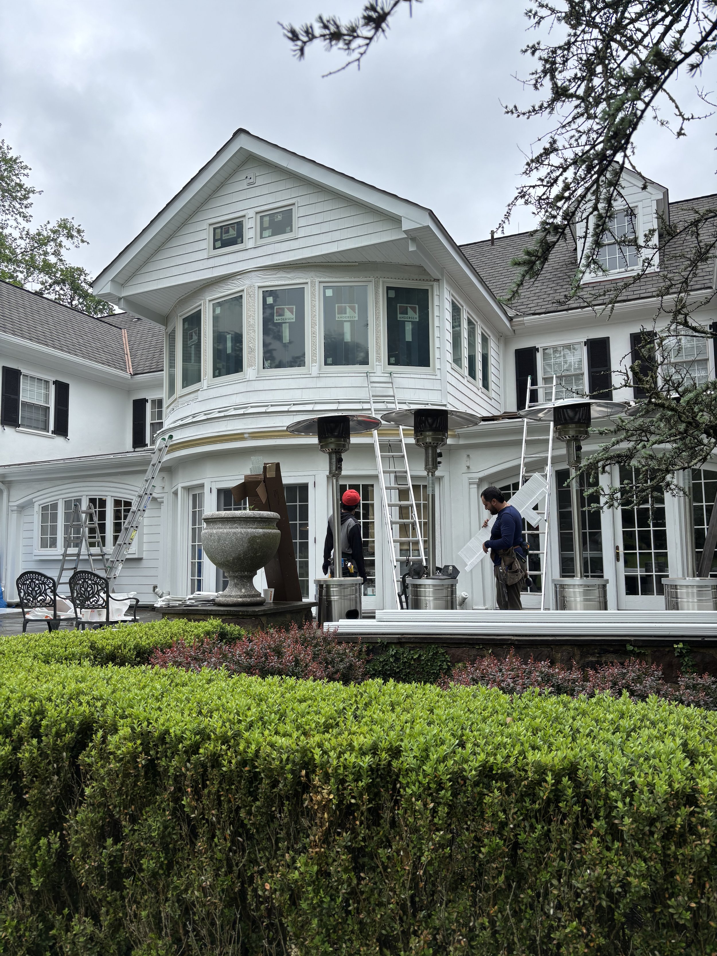 Construction workers working on the exterior of a large white house, installing or repairing a deck or balcony, with ladders and propane heaters in front, and green bushes in the foreground.