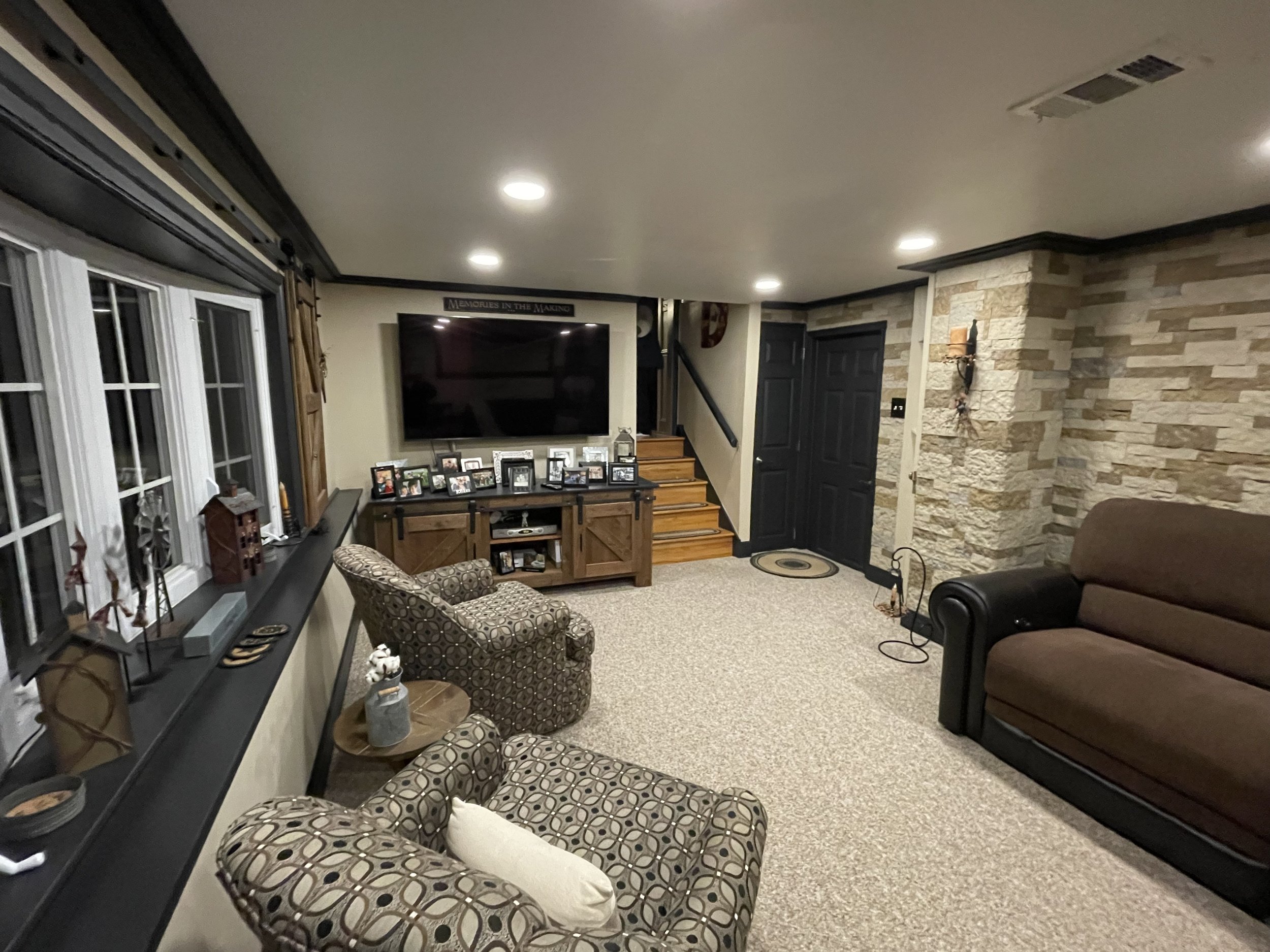 Living room with a large wall-mounted TV, framed photos on a wooden TV stand, patterned armchair, brown sofa, stone accent wall, window with decorations, and ceiling lights.