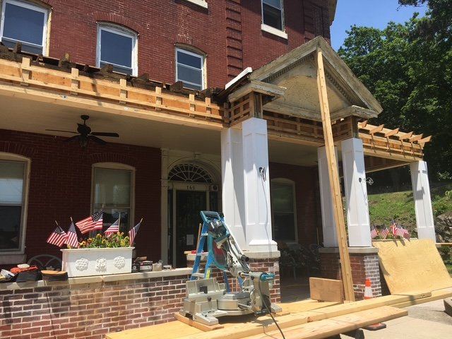 Construction work on a brick building's porch, with wooden framing, construction tools, and American flags on a flower box in front.