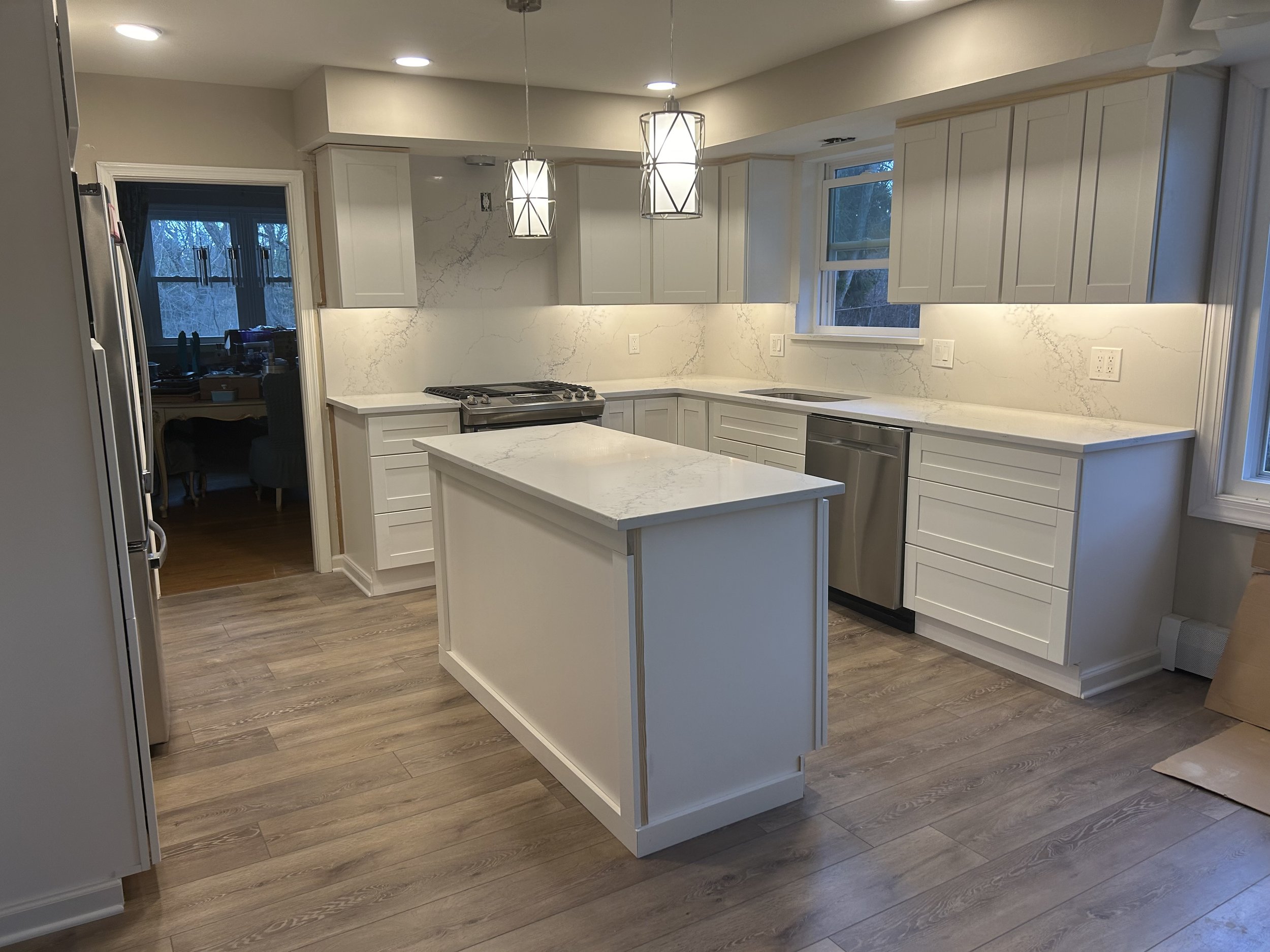 Modern kitchen with white cabinets, marble countertops, a small island, stainless steel appliances, and light wood flooring.