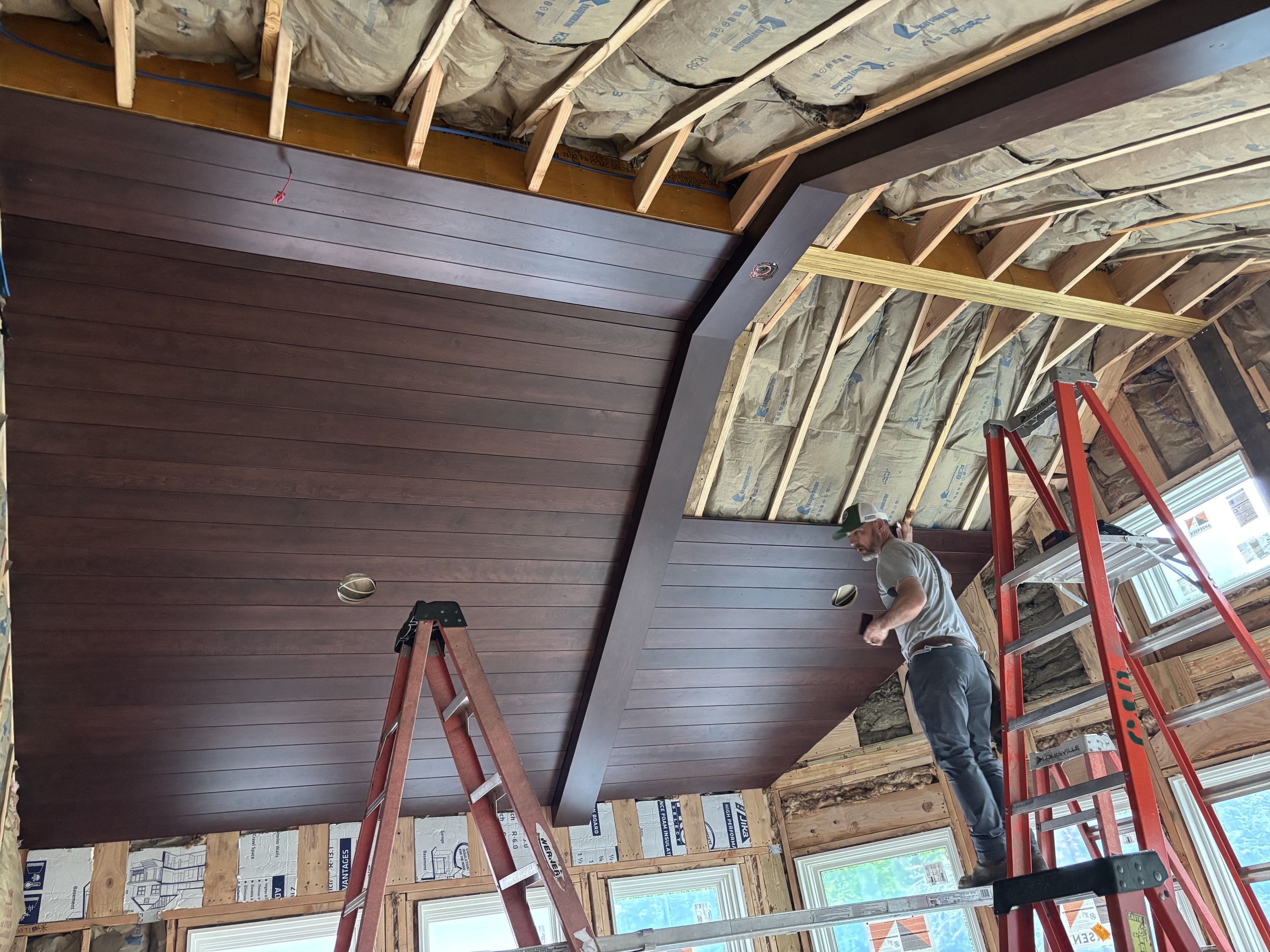 A man working on installing a dark wood-paneled ceiling in a room under construction. He is standing on a red ladder, with windows and insulation visible in the background.