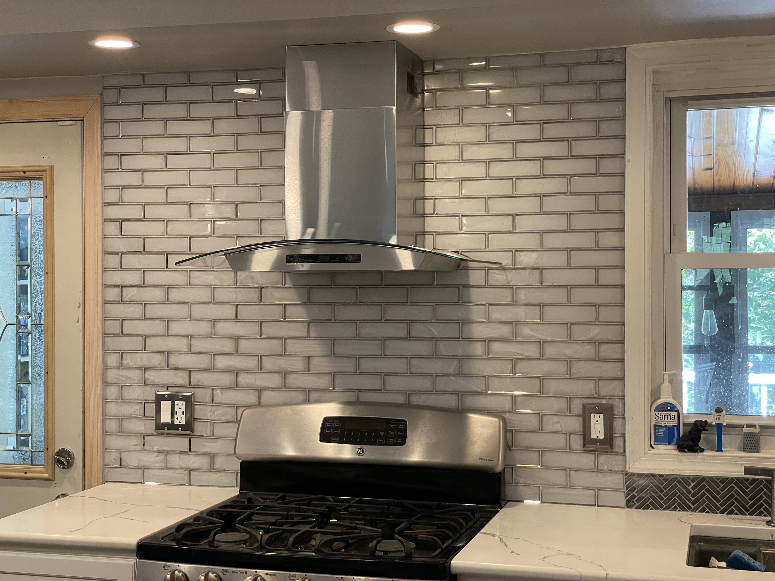 Kitchen with white marble countertop, stainless steel gas stove, stainless steel range hood, and gray brick backsplash. There is a window on the right with cleaning supplies on the windowsill, and a door on the left with glass panels.