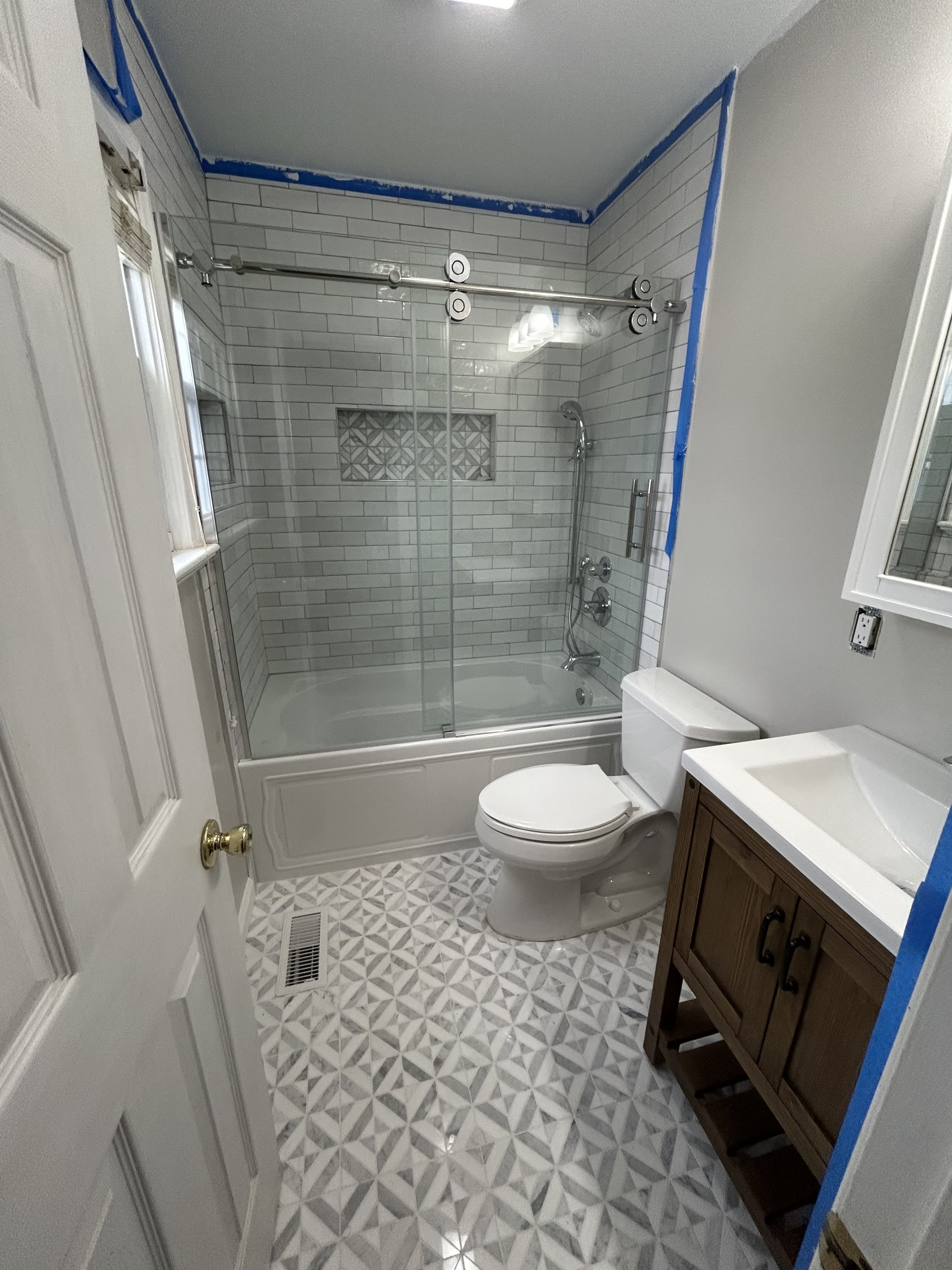 Bathroom with walk-in shower, toilet, and wooden vanity with a white sink. The walls and floor feature patterned tile, with blue painter's tape along the ceiling and edges.