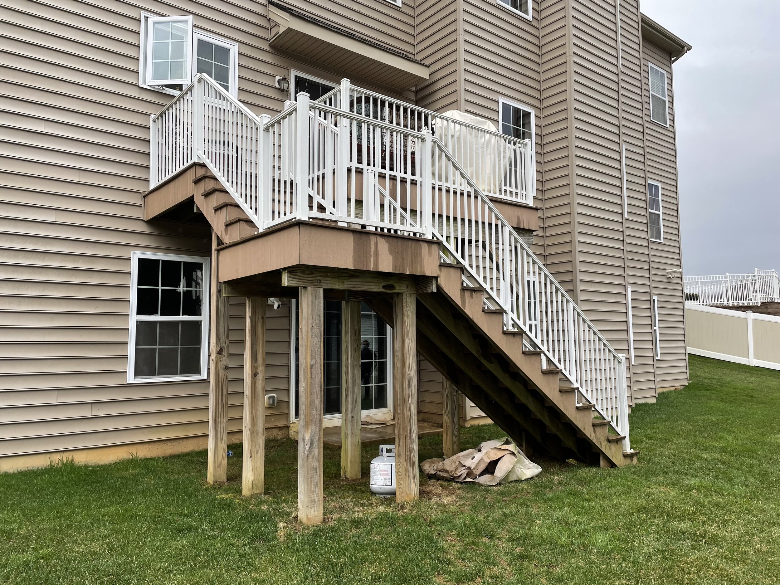 Backyard view of a beige house with an updated wooden deck and white railing, supported by four wooden posts, with stairs leading down to a grassy yard.