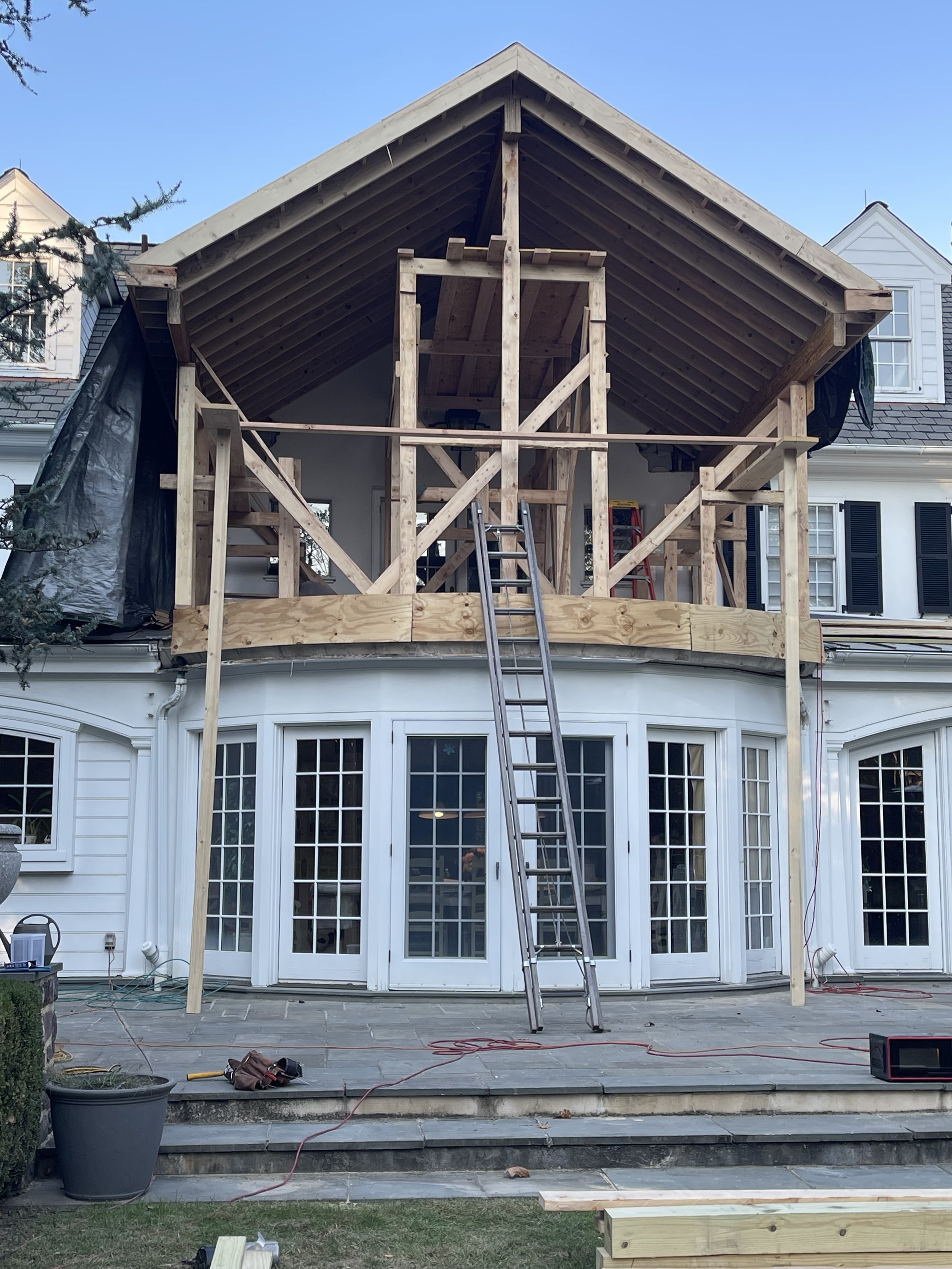 House undergoing construction with a new wooden upper-level deck and roof framing, ladders, construction tools, and materials present.