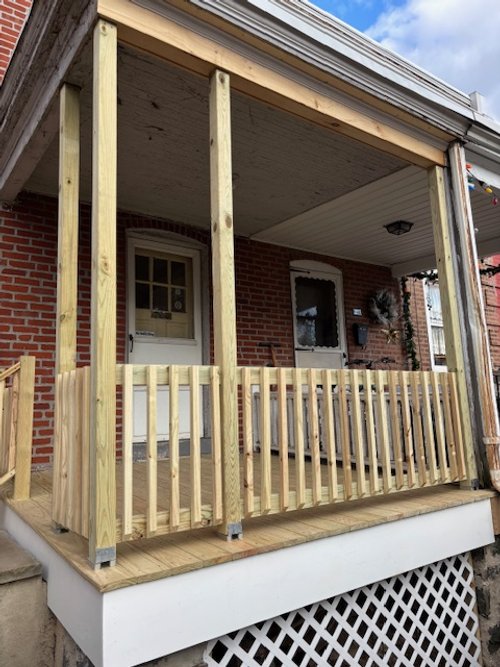 Front porch under construction with new wooden railing and posts, brick house with a white door and window, partly cloudy sky.