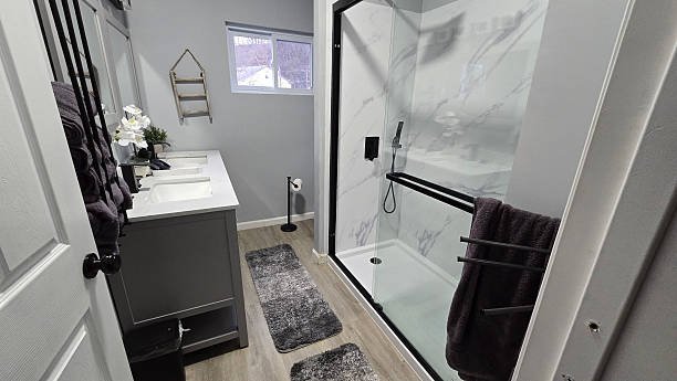 Modern bathroom with a gray vanity, white countertop, black fixtures, a glass-enclosed shower with marble walls, and two gray rugs on light wooden floor.