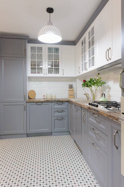 Modern kitchen with gray lower cabinets, white upper cabinets with glass doors, tiled backsplash, and patterned tile floor. A white pendant light hangs from the ceiling.