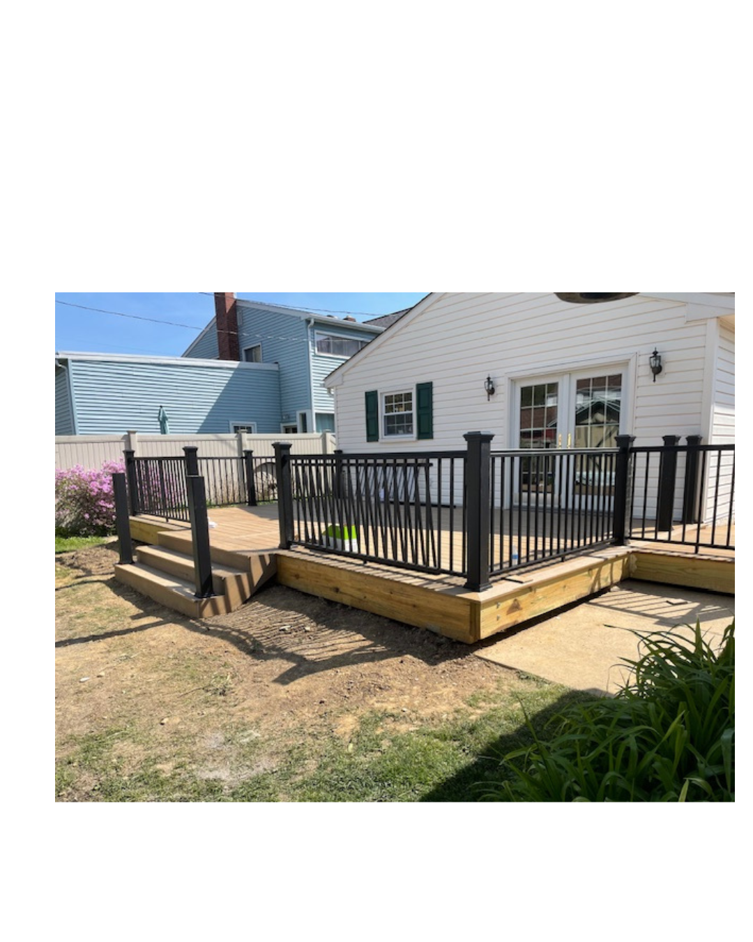 Wooden deck with black metal railing attached to a white house with green shutters, with steps leading down to a dirt yard and neighboring houses in the background.
