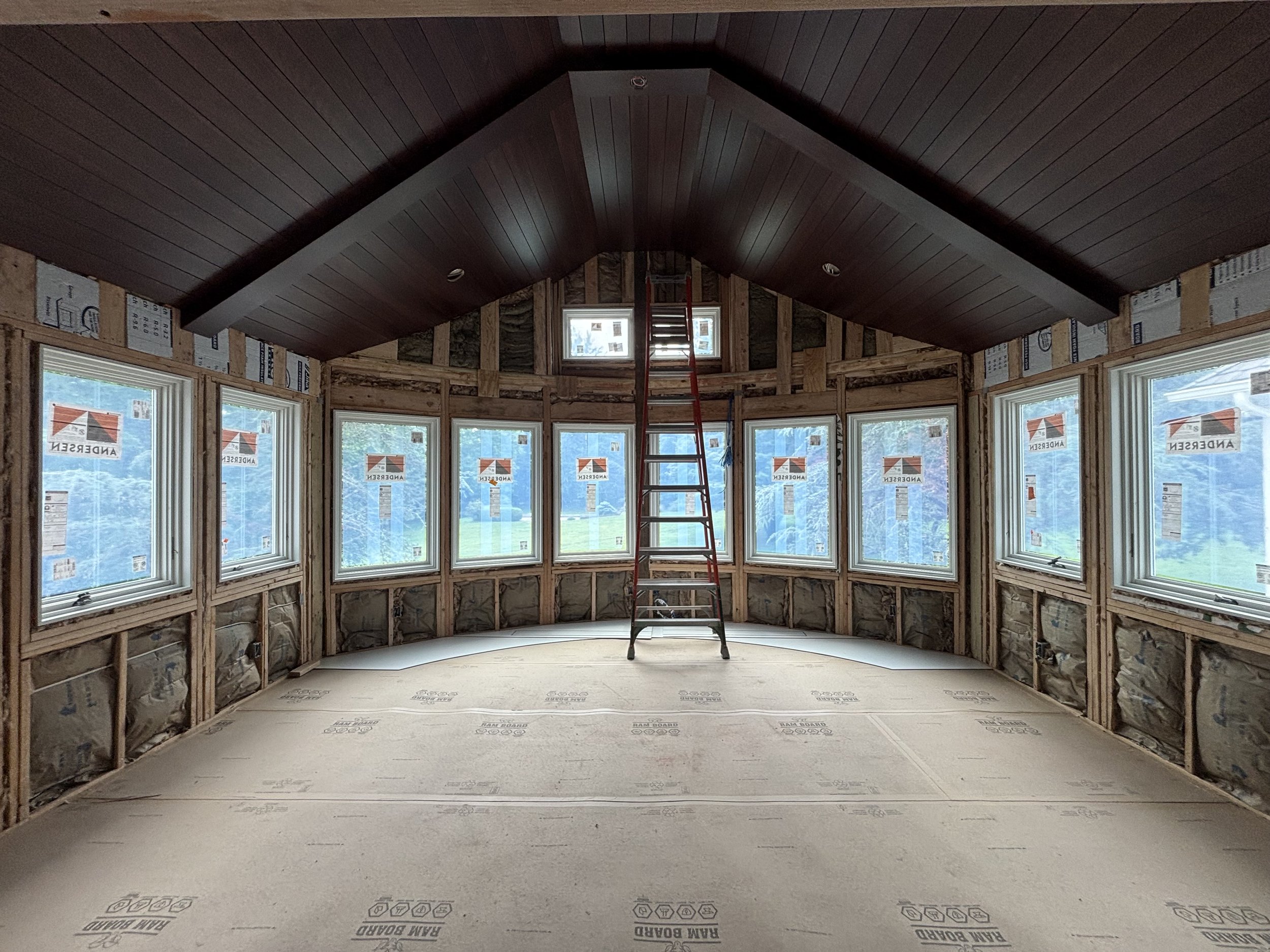 Interior of a house under construction with large bay windows, a vaulted ceiling with dark wood paneling, and insulation visible around the frames. A red ladder is positioned in the center.