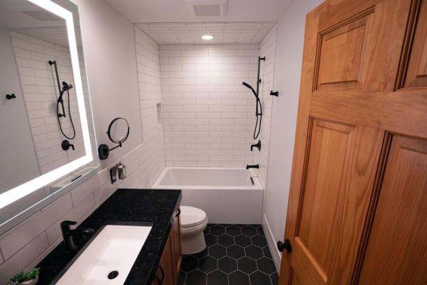 Modern bathroom with white subway tile walls, black fixtures, a bathtub with dual showerheads, a black countertop with a white sink, a mirror with LED lighting, a wooden cabinet, and a textured black tile floor.