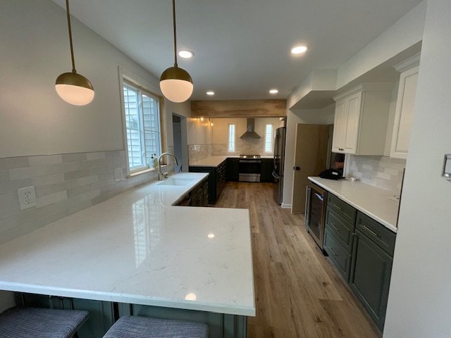Modern kitchen with white countertops, white and green cabinets, wooden flooring, two pendant lights, and a view into the adjacent stove area with stainless steel appliances.