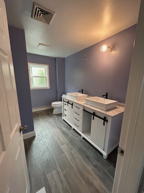 Bathroom with gray walls, a white vanity with two vessel sinks, and a toilet near a window.