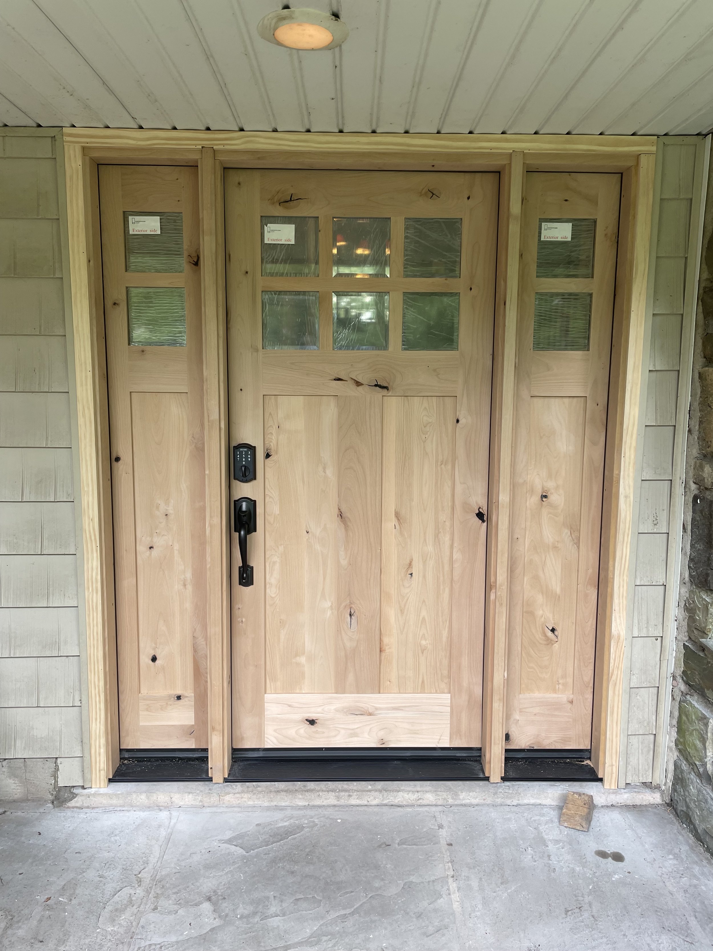 A wooden front door with glass panels surrounded by matching wood trim and an electrical lock, set within a porch with a concrete floor and ceiling.