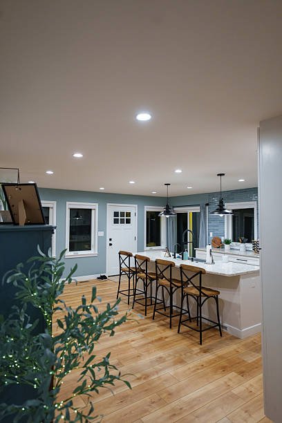 Modern kitchen and dining area with a light blue wall, white cabinets, a kitchen island with barstools, pendant lighting, and hardwood flooring.