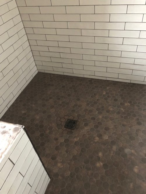 Corner of a bathroom with white subway tile walls and a dark hexagonal tile floor featuring a drain.