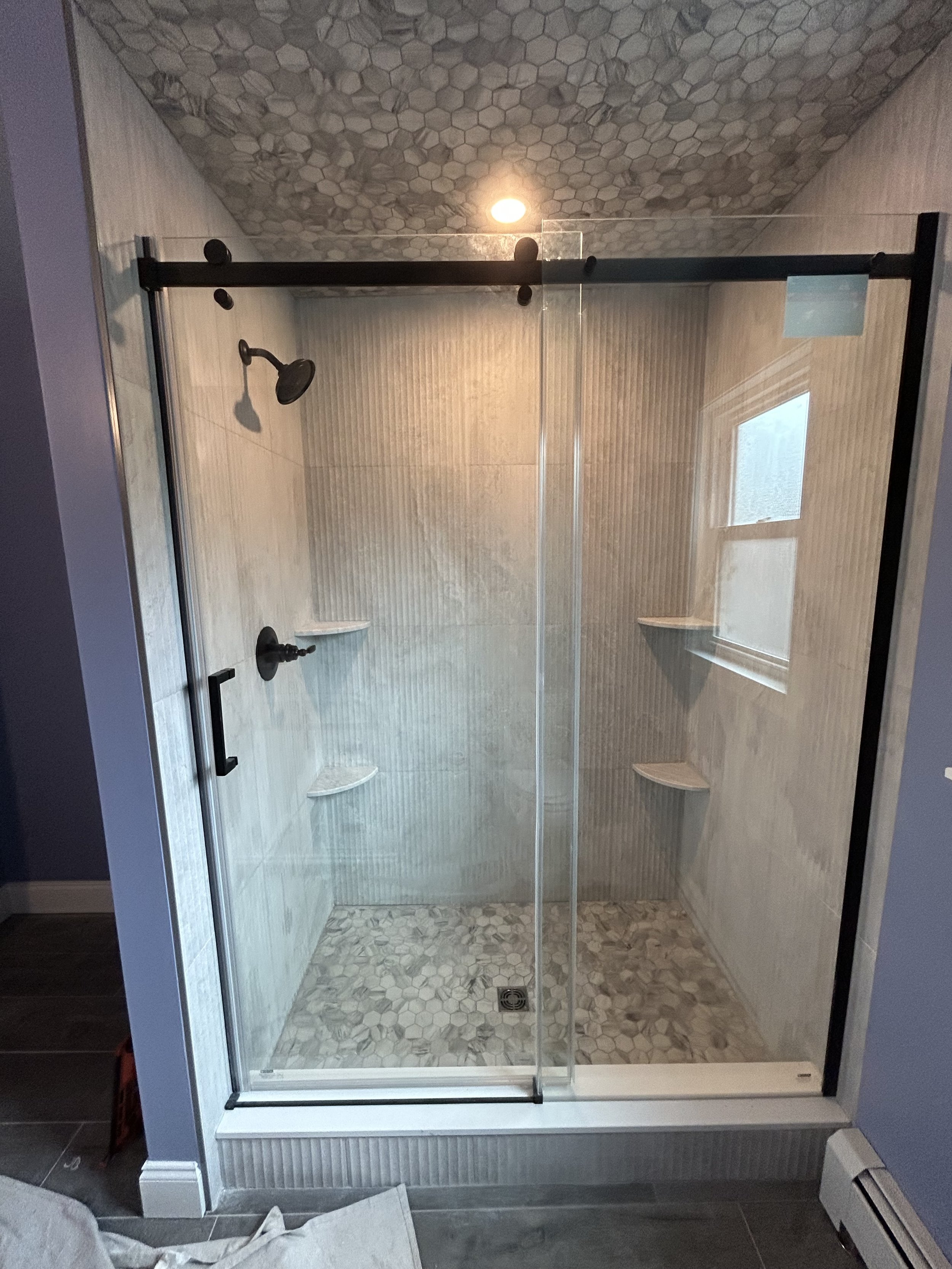 A modern glass-enclosed shower with black hardware, stone-tile flooring, and a small window, located in a bathroom with gray and white decor.
