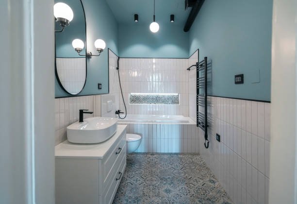 Modern bathroom with light blue walls, white tiled lower walls, a bathtub, toilet, white vanity with a vessel sink, oval mirror, wall-mounted light fixtures, black fixtures, and patterned floor tiles.