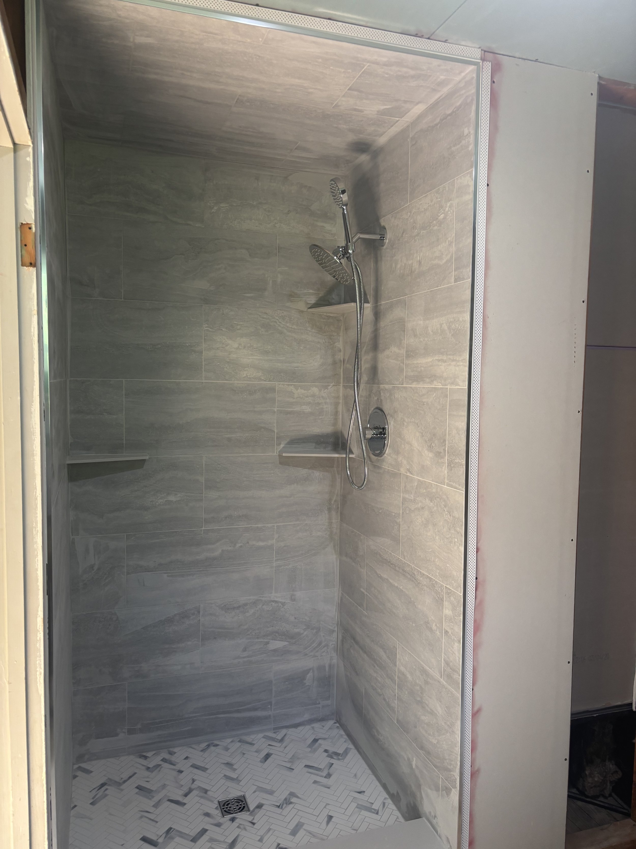 A tiled walk-in shower with gray stone tiles, a modern silver showerhead, and two built-in corner shelves.