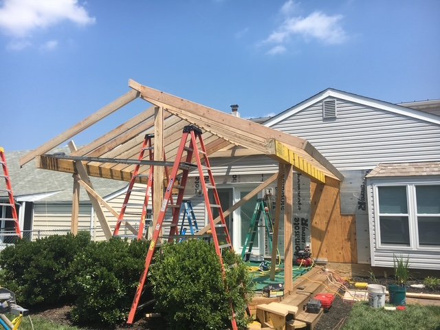 Construction site of a house roof with wooden beams, ladders, and construction tools, with surrounding bushes and a neighboring house in the background.