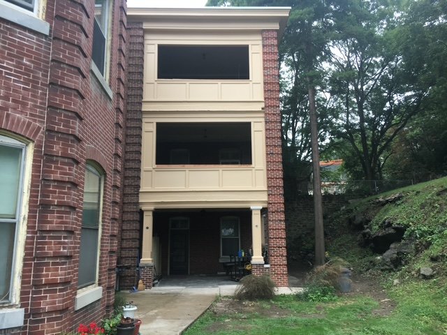 Front of a three-story house with a brick facade and beige siding on the upper two levels, with an outdoor sitting area on the ground level, surrounded by greenery and trees.
