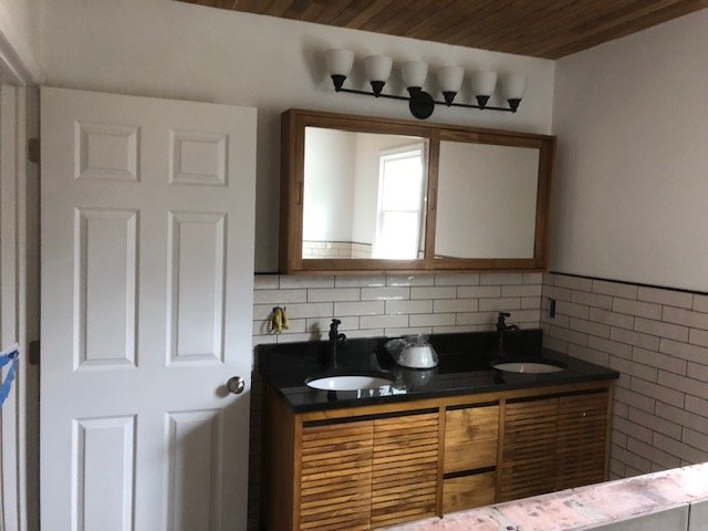 Bathroom vanity with dual sinks, black countertop, wooden cabinet, large mirror, and white wall with subway tile backsplash.