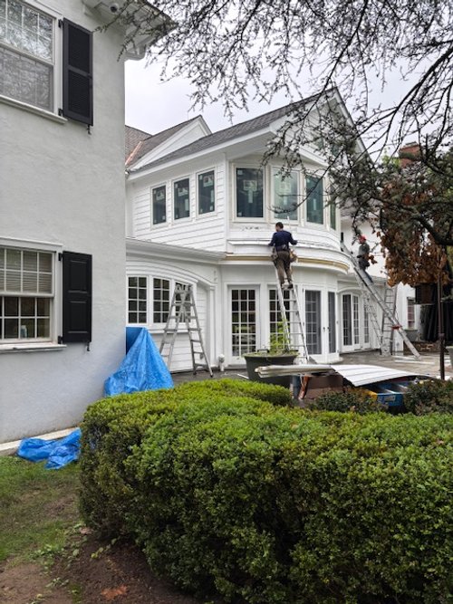 Workers painting the exterior of a white two-story house with tall windows, using ladders and scaffolding, in a yard with bushes and trees.