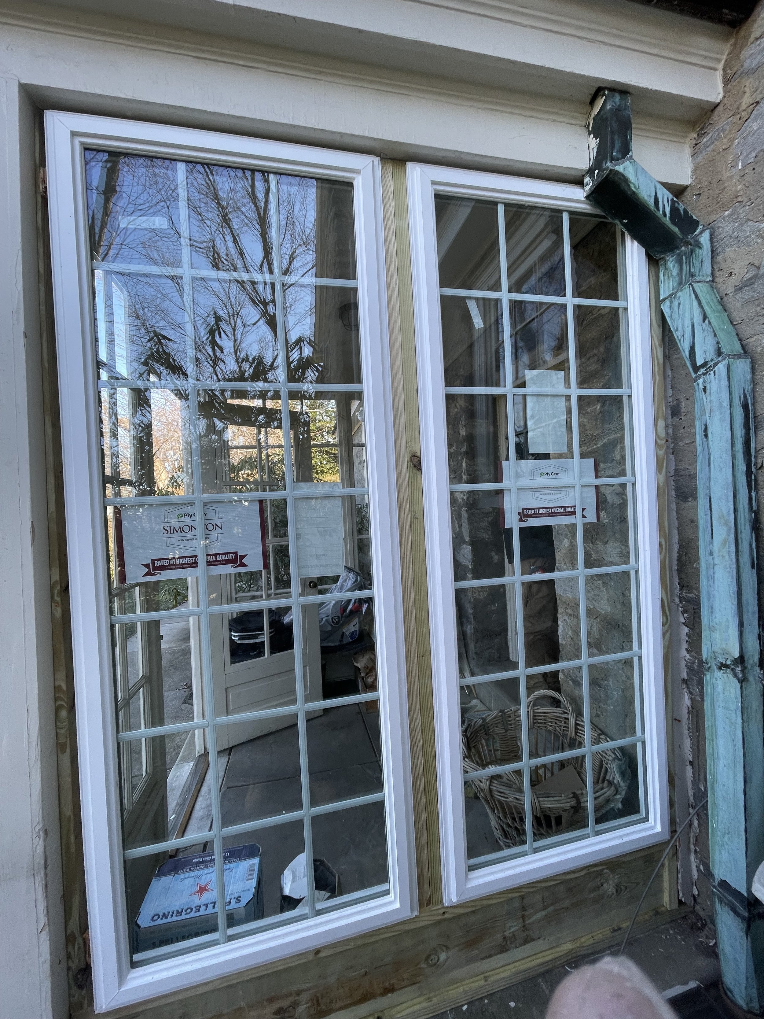 A pair of white-frame double-hung windows installed on a house with visible wooden framing and stone wall. The windows reflect trees and sky outside, and there are construction supplies inside.