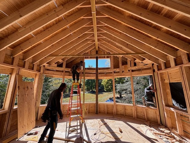 Interior view of a house under construction with wooden framing and workers installing the roof.