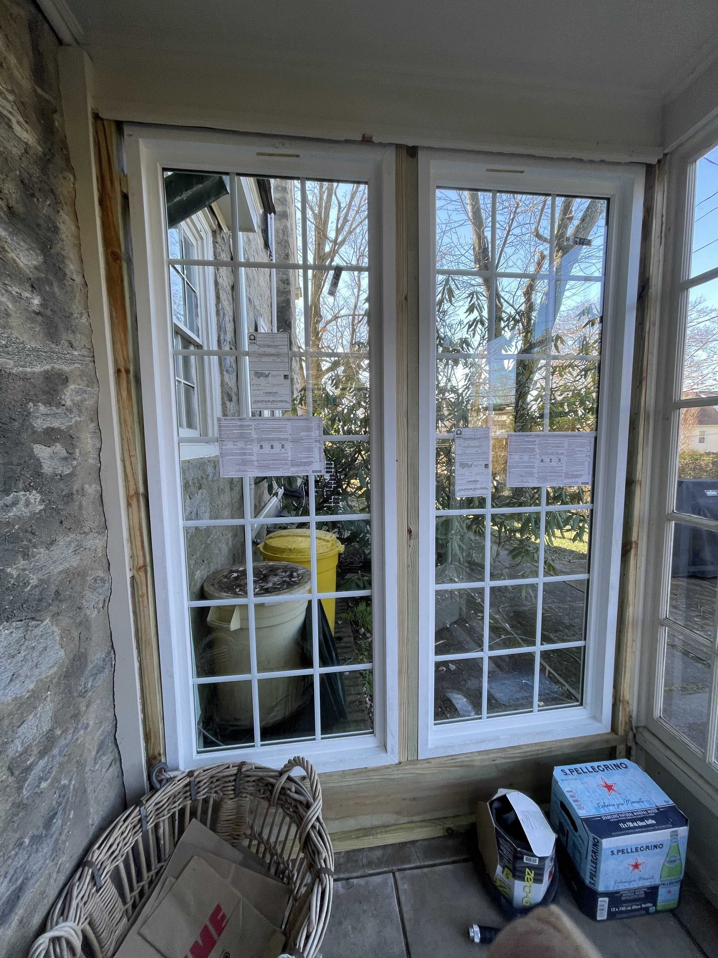 Interior of a porch or sunroom showing new white-framed double glass doors leading outside, with construction or renovation materials on the floor.