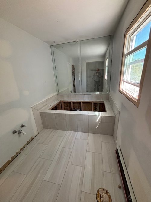 Bathroom with an empty bathtub frame, a mirror on the back wall, a window on the right, and some construction materials on the floor.