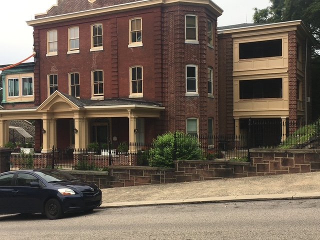 Old brick building with a beige porch and a modern beige addition on the side, parked black car in front, street and sidewalk in foreground.