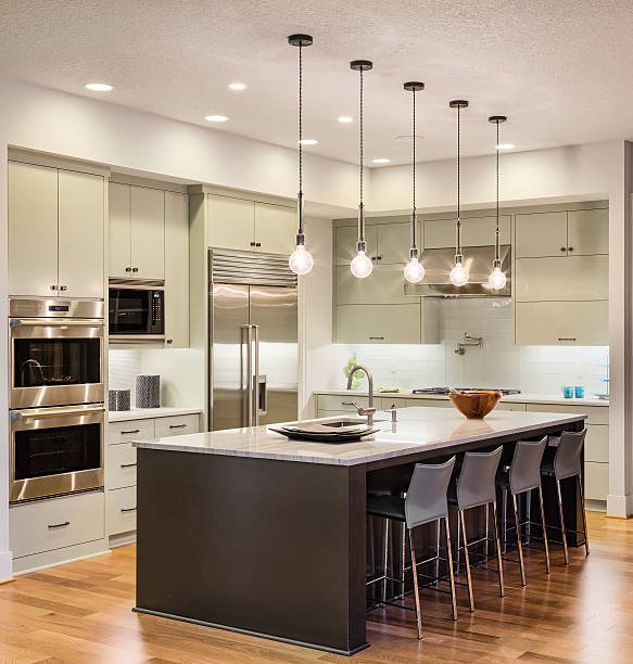 Modern kitchen featuring light green cabinetry, stainless steel appliances including a double oven and refrigerator, a large island with a dark base and white countertop, five black bar stools, pendant lighting, and hardwood floors.