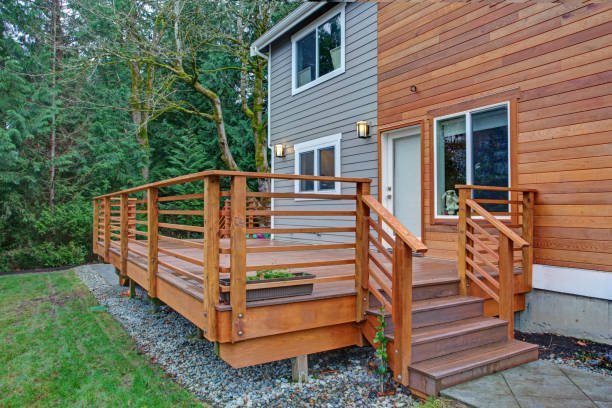 Wooden ramp and stairs attached to the side of a house with a gray and wooden exterior, overlooking a green backyard with trees.