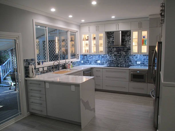 Modern kitchen with white cabinets, gray countertops, and stone backsplash under ceiling lights, with a sliding glass door leading outside.