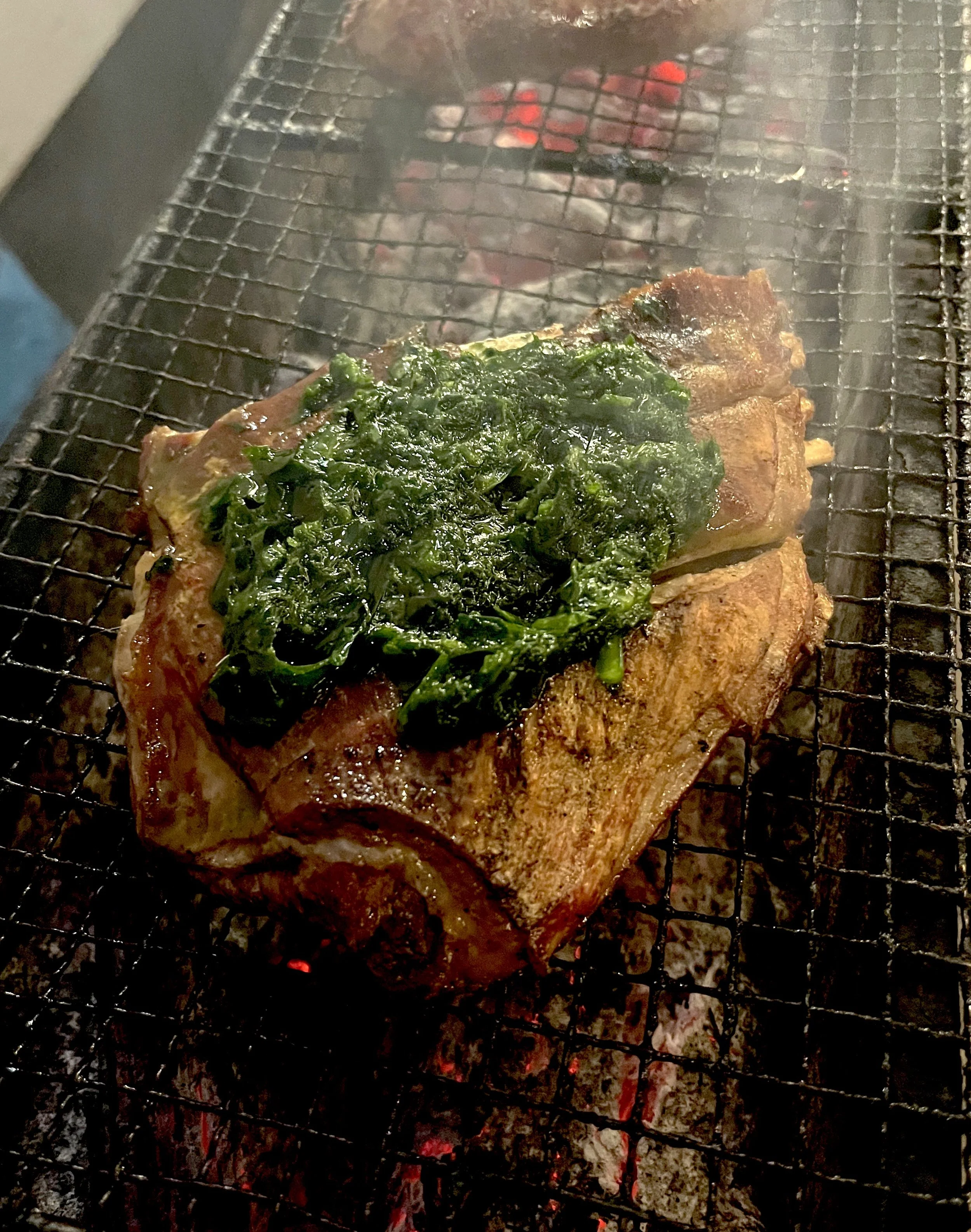 A piece of steak topped with chopped cooked greens, grilling on a barbecue grill with red hot coals underneath.