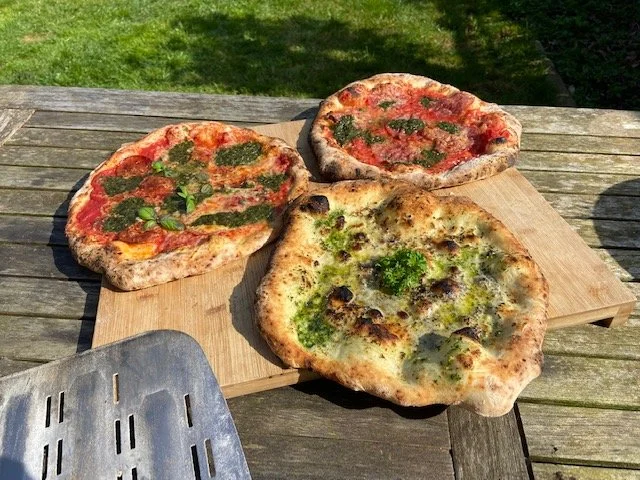 Three pizzas, two Margherita and one with green herbs and mushrooms, on a wooden cutting board outdoors.