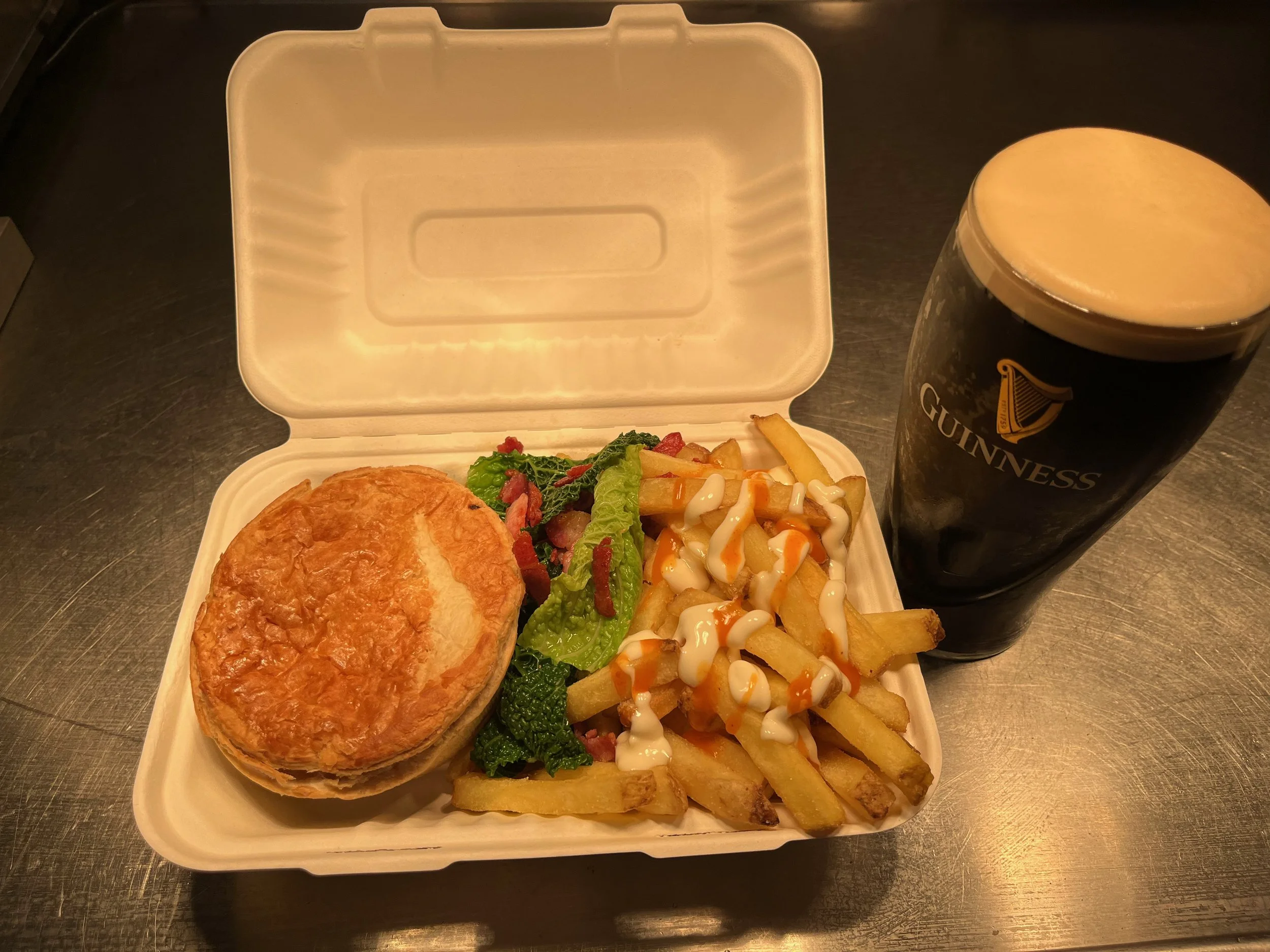 A takeout container with a meat pie, mixed vegetables, and French fries topped with mayonnaise and spicy sauce, alongside a pint of Guinness beer.