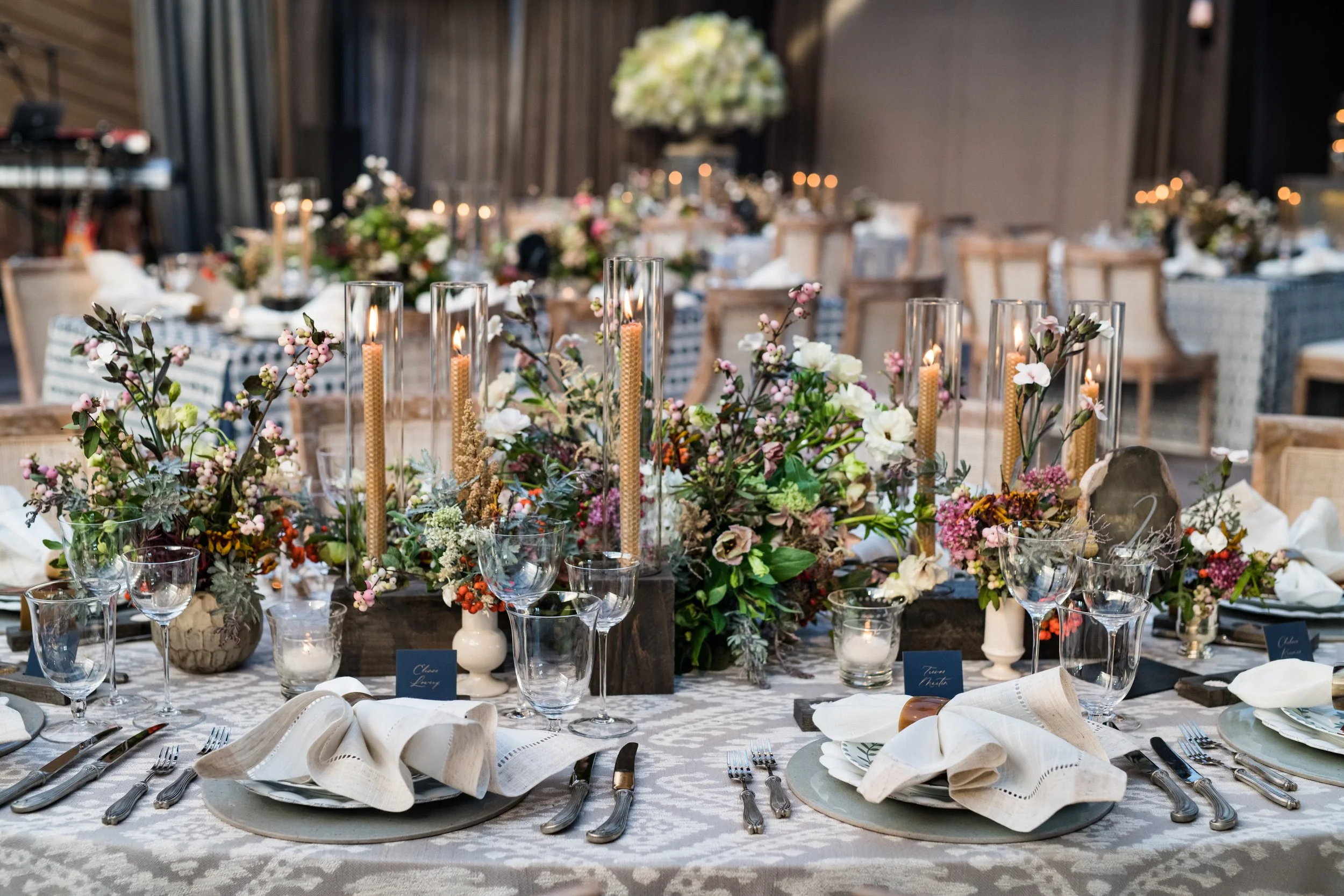 A decorated banquet table with floral centerpieces, candles in glass holders, and place settings including plates, silverware, and folded napkins at an elegant event.
